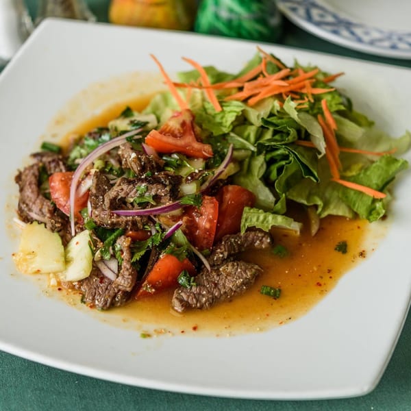 Thai beef salad with tomatoes, onions, cucumbers, and lettuce on a white plate.